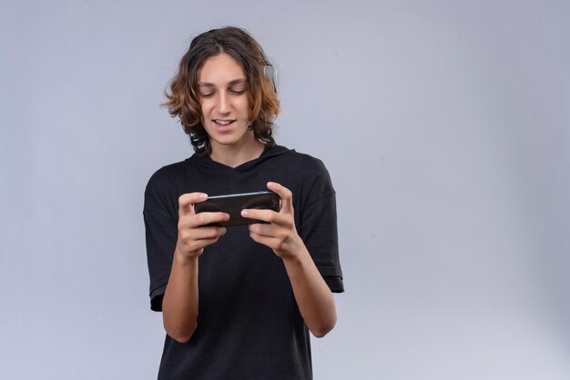 Ragazzo sorridente con capelli lunghi in maglietta nera che gioca gioco da un telefono sul muro bianco