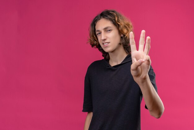 Улыбающийся парень с длинными волосами в черной футболке показывает троих пальцами на розовой стене