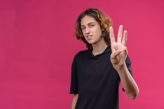 Smiling guy with long hair in black t-shirt showimg three with his fingers on pink wall