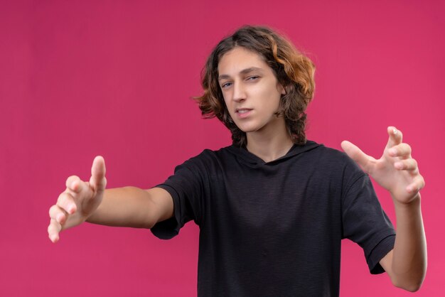 Smiling guy with long hair in black t-shirt showing size on pink wall