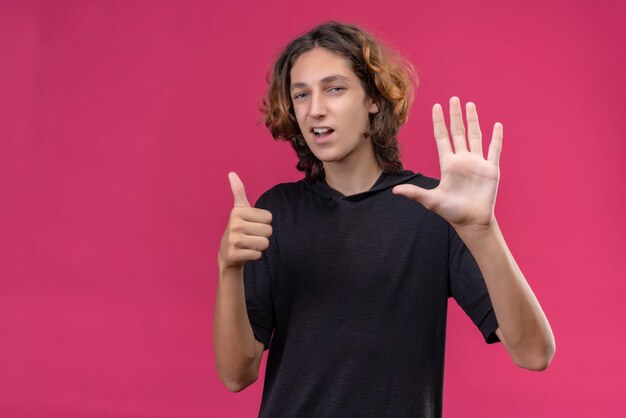 Smiling guy with long hair in black t-shirt shows thumb up on pink wall