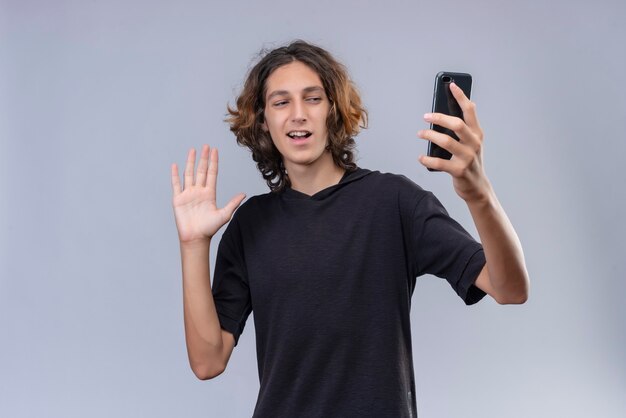 Улыбающийся парень с длинными волосами в черной футболке разговаривает по телефону на белой стене