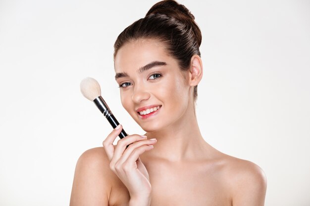 smiling half-naked woman with fresh skin holding brush for makeup close to face applying concealer