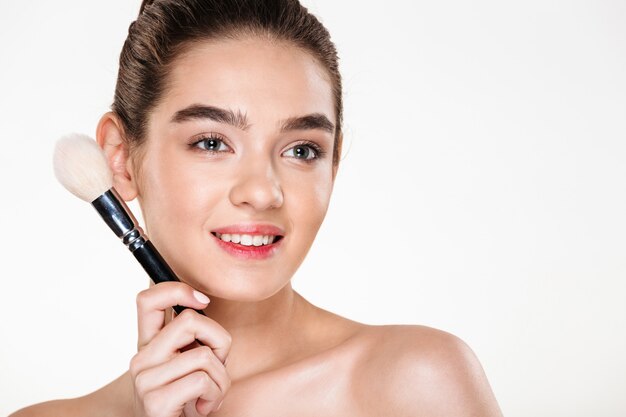 smiling half-naked woman with fresh skin holding brush for makeup close to face and looking aside