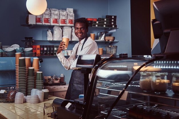 제복을 입은 잘생긴 아프리카계 미국인 바리스타가 커피숍에 서서 커피 한 잔을 들고 웃고 있습니다.