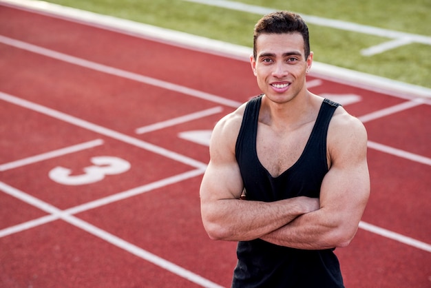 Foto gratuita l'atleta bello sorridente in un'attrezzatura sportiva con le sue armi ha attraversato sulla pista di corsa che esamina la macchina fotografica