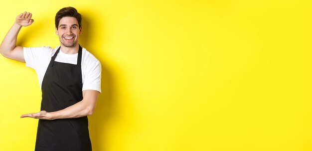 Free photo smiling handsome barista showing something long or large standing over yellow background
