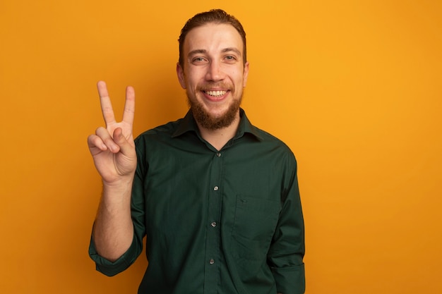 Smiling handsome blonde man gestures victory hand sign isolated on orange wall