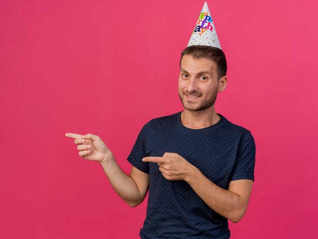 コピースペースとピンクの背景に分離された両手で横を指している誕生日の帽子をかぶってハンサムな白人男性の笑顔