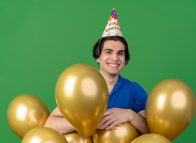 Smiling handsome caucasian man wearing birthday cap standing with helium balloons 