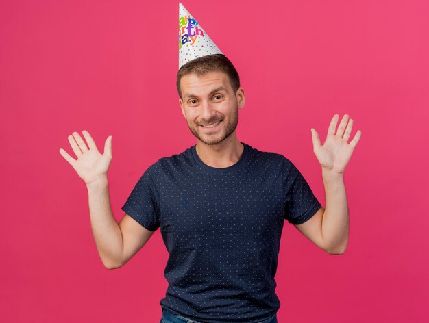 Smiling handsome caucasian man wearing birthday cap stands with raised hands isolated on pink background with copy space