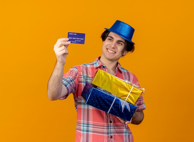 Smiling handsome caucasian man wearing blue party hat holds gift boxes and looks at credit card 