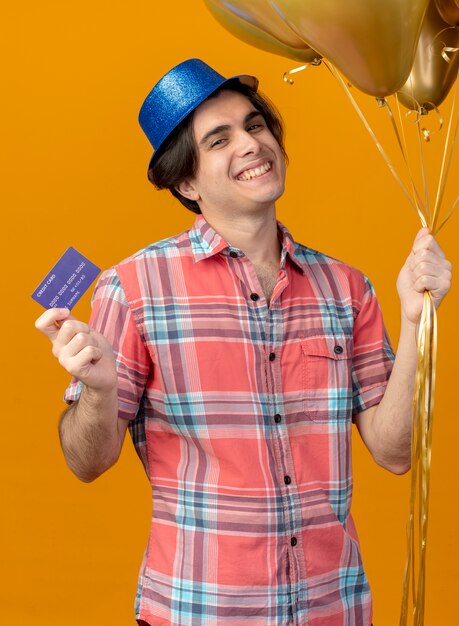 青いパーティー ハットをかぶった笑顔のハンサムな白人男性が、カメラを見てヘリウム風船とクレジット カードを保持しています。