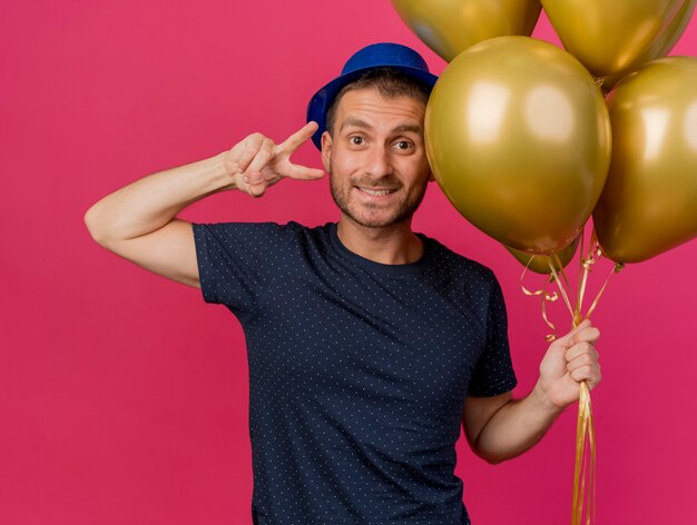 Smiling handsome caucasian man wearing blue party hat holds helium balloons and gestures victory hand sign isolated on pink background with copy space