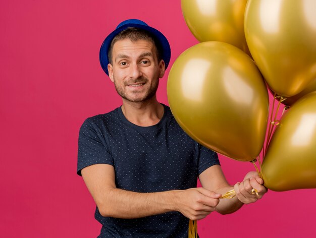 Smiling handsome caucasian man wearing blue party hat holds helium balloons isolated on pink background with copy space