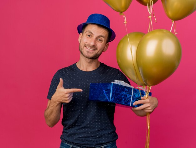 Smiling handsome caucasian man wearing blue party hat holds and points at helium balloons isolated on pink background with copy space