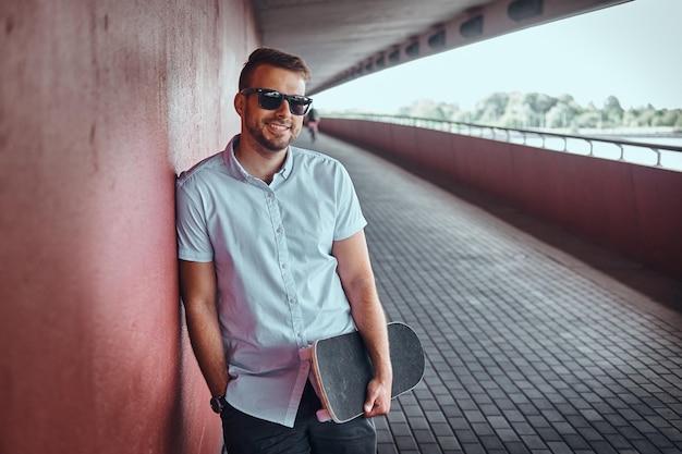 Free photo smiling handsome fashionable guy in sunglasses dressed in a white shirt and shorts holds a skateboard,  leaning against a wall while standing on a footpath under the bridge.
