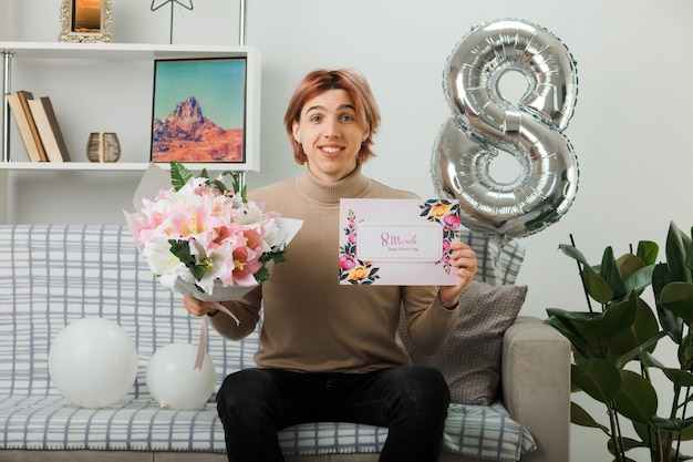 Smiling handsome guy on happy women day holding bouquet with greeting card sitting on sofa in living room