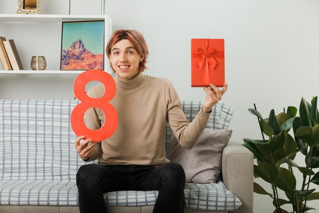 Smiling handsome guy on happy women day holding number eight with present sitting on sofa in living room
