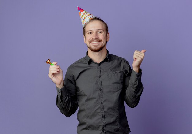 Smiling handsome man in birthday cap holds whistle and thumbs up isolated on purple wall