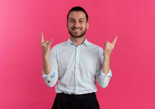 Smiling handsome man gestures horns hand sign isolated on pink wall