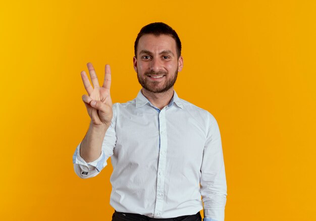Smiling handsome man gestures three with fingers isolated on orange wall