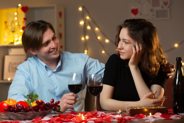 Улыбающийся красавец держит бокал вина и смотрит на довольную красивую женщину, сидящую за столом в гостиной в день святого валентина