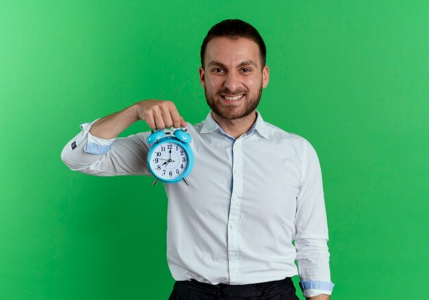 Foto gratuita l'uomo bello sorridente tiene la sveglia che sembra isolata sulla parete verde