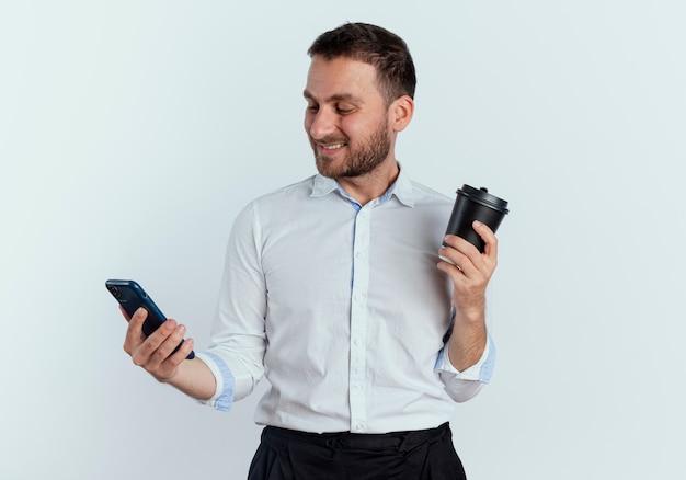 Smiling handsome man holds coffee cup and looks at phone isolated on white wall