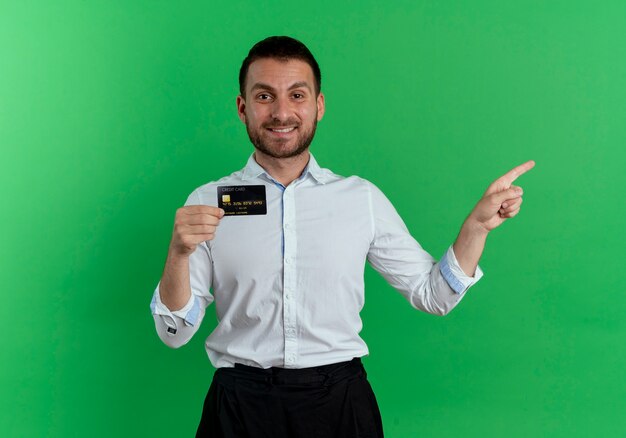 Smiling handsome man holds credit card and points at side isolated on green wall