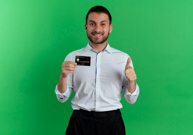 Smiling handsome man holds credit card and thumbs up isolated on green wall