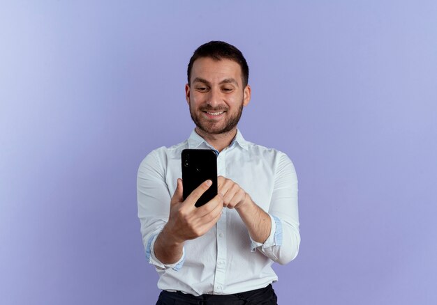 Улыбающийся красавец держит и смотрит на телефон, изолированный на фиолетовой стене