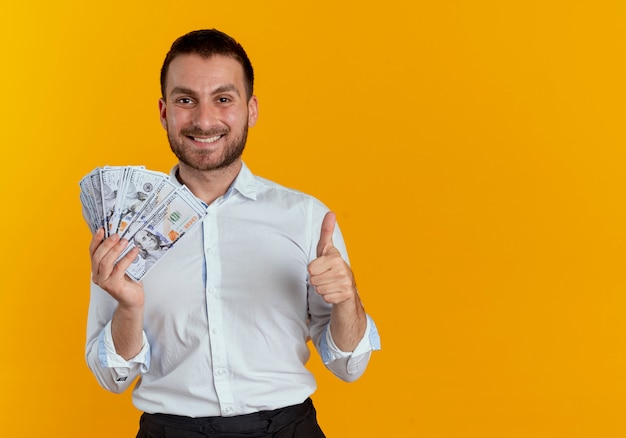 Smiling handsome man holds money and thumbs up isolated on orange wall