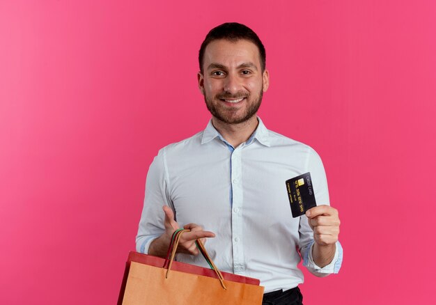 Smiling handsome man holds paper shopping bags and credit card isolated on pink wall