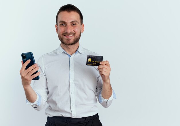 Smiling handsome man holds phone and credit card isolated on white wall