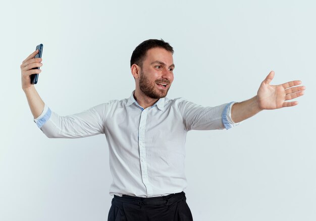 Smiling handsome man holds phone and points at side with hand isolated on white wall