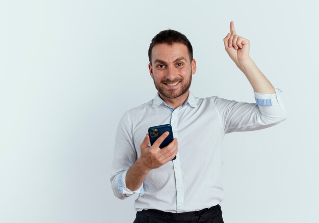 Smiling handsome man holds phone and points up isolated on white wall