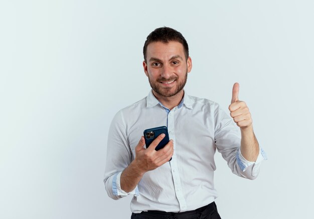 Smiling handsome man holds phone and thumbs up looking isolated on white wall