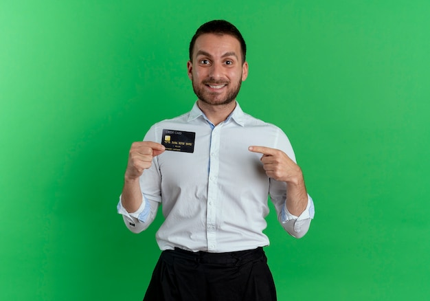 Smiling handsome man holds and points at credit card isolated on green wall