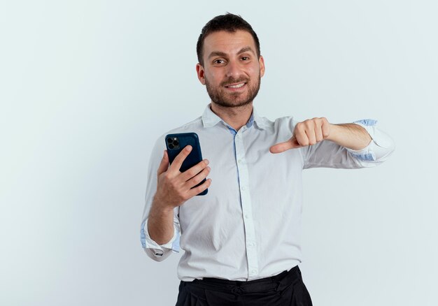 Smiling handsome man holds and points at phone looking isolated on white wall