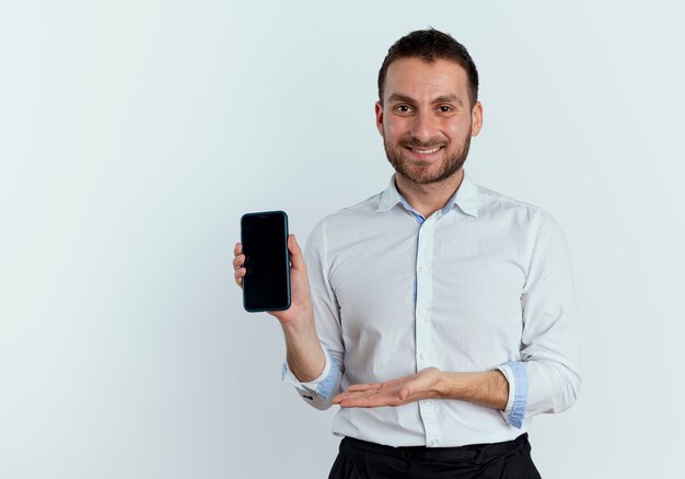 Smiling handsome man holds and points at phone with hand isolated on white wall