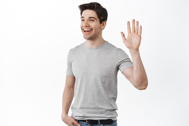 Smiling handsome man looking and waving aside, say hi to a friend, casually standing in relaxed pose against white wall