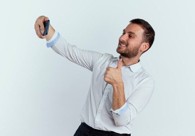 Smiling handsome man looks at phone and thumbs up isolated on white wall
