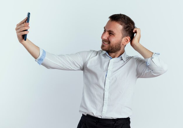 Smiling handsome man puts hand on head behind looking at phone isolated on white wall