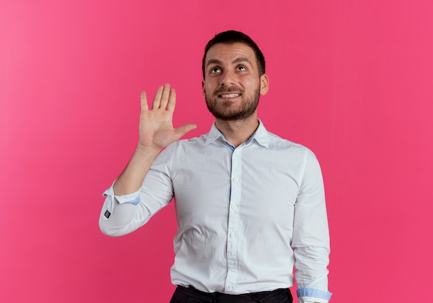 Smiling handsome man raises hand looking up isolated on pink wall