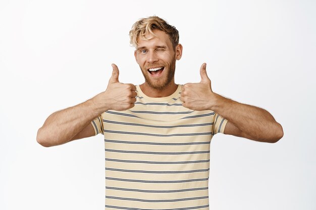 Smiling handsome man showing thumbs up and winking say yes praise and compliment standing in tshirt over white background