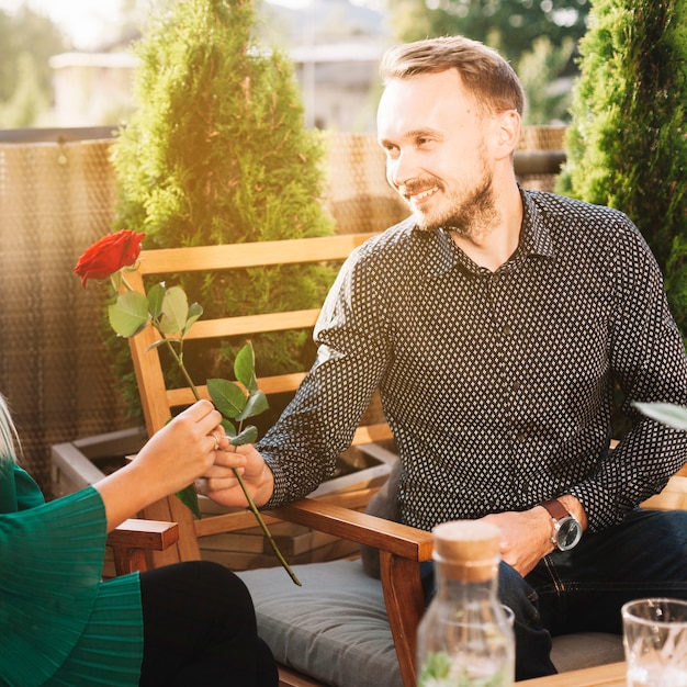 Smiling handsome man sitting on chair giving red rose to her girlfriend