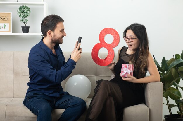 3月の国際女性の日にリビングルームのソファに座っている赤い8の数字とギフトボックスを保持している光学メガネでうれしそうなきれいな若い女性の写真を撮るハンサムな男性の笑顔