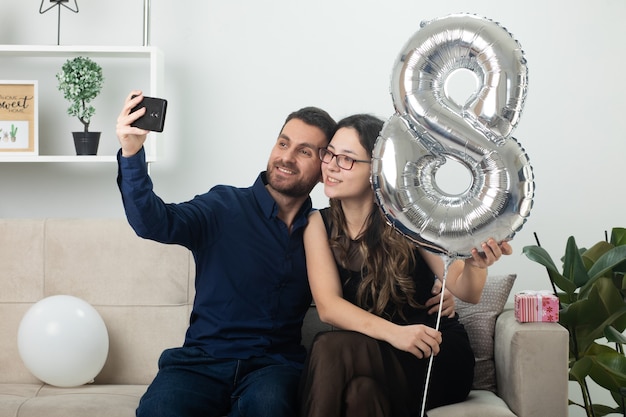 Улыбающийся красавец делает селфи по телефону с симпатичной молодой женщиной в оптических очках, держащей воздушный шар в форме восьми и сидящей на диване в гостиной в международный женский день в марте