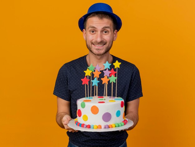 Smiling handsome man wearing blue party hat holds birthday cake isolated on orange wall with copy space